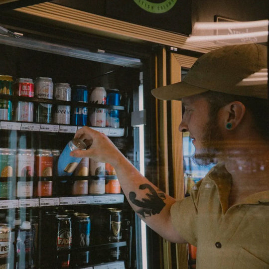 A hand hold a can of Small Gods Neverending German Pilsner in front of a fridge full of beer.